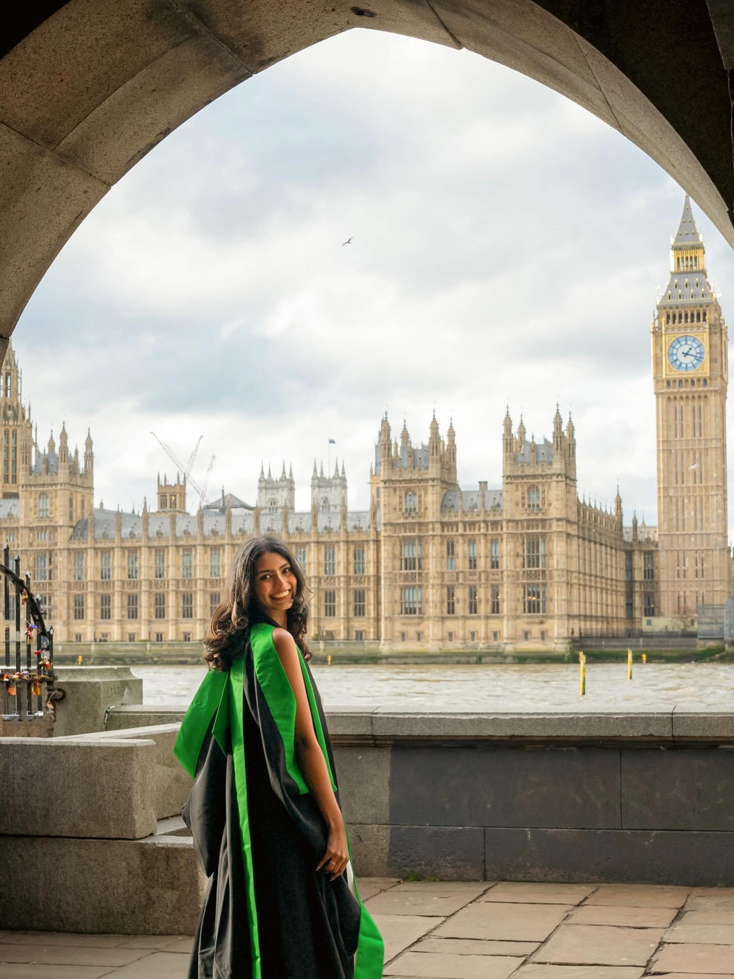 Graduate in cap and gown photographed under an archway near Big Ben and the Houses of Parliament, London
